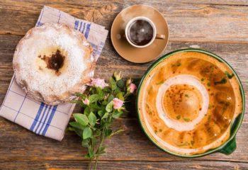 A Gugelhupf with its ceramic baking dish and a cup of coffee and a field flowers birthday bouquet on a wooden table with a wooden background.